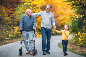 Elderly man with walker walking alongside adult son and young grand son on an autumnal walk.